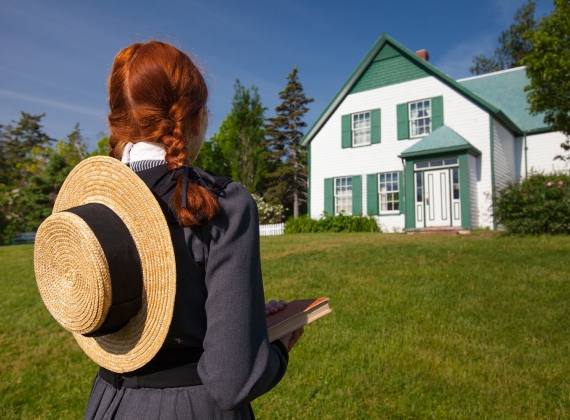 A girl with red hair in braids and a sun hat on her back looking at a white house on a sunny day.