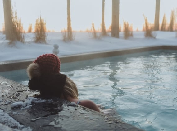 Two people wearing winter hats watching a sunset in a hot tub on a snowy evening