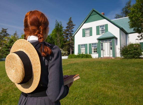 A girl with red hair in braids and a sun hat on her back looking at a white house on a sunny day.