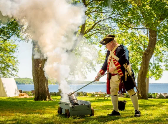 A man dressed in traditional privateer costume laying coals on a fire in a sunny park.