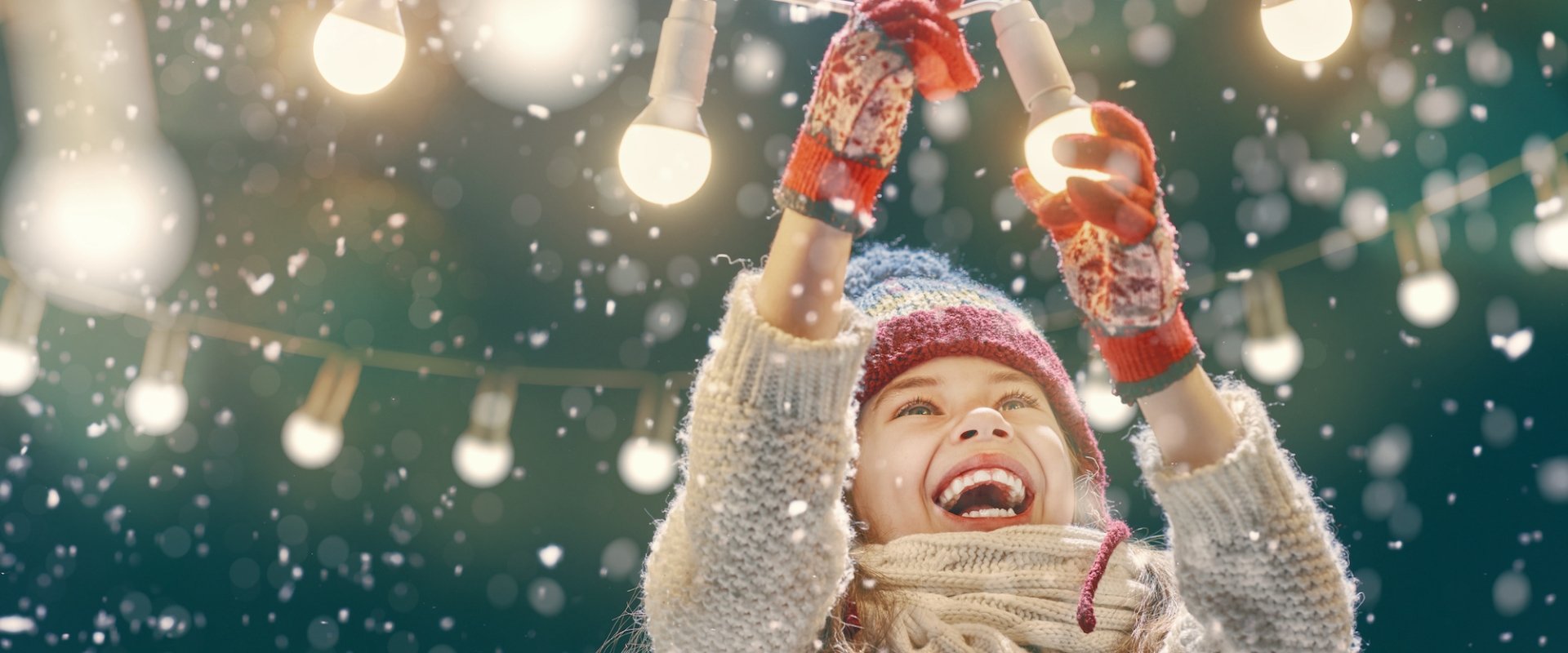 Child smiling and playing with string globe lights in a light snowfall.
